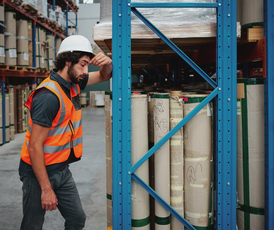 Warehouse Worker wearing a white helmet and orange hi-vis top looking stressed