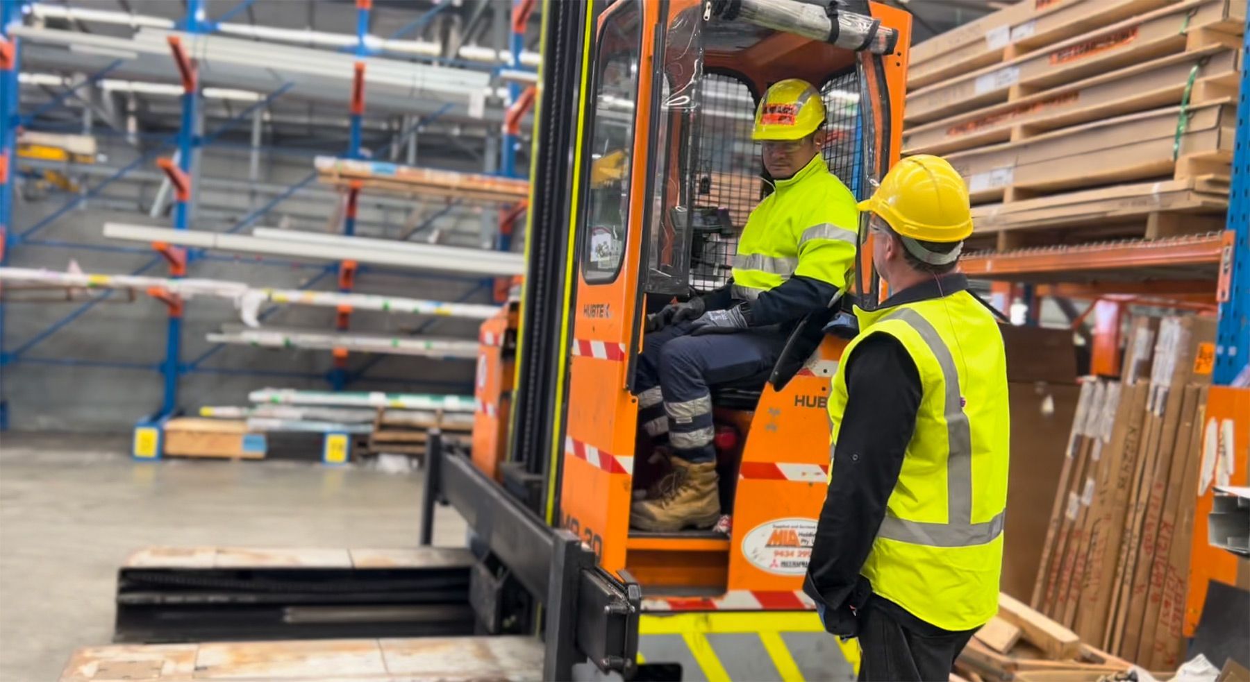 A man wearing yellow hi-vis and a yellow hard hat in a warehouse talking to another man in safety gear who is on an orange forklift 