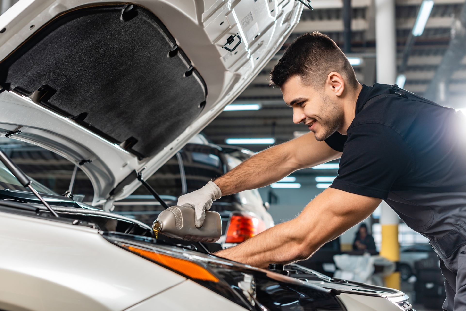 Mechanic pouring oil into a car engine, smiling. White car in a garage. Mechanic pouring oil into a car engine, smiling. White car in a garage.