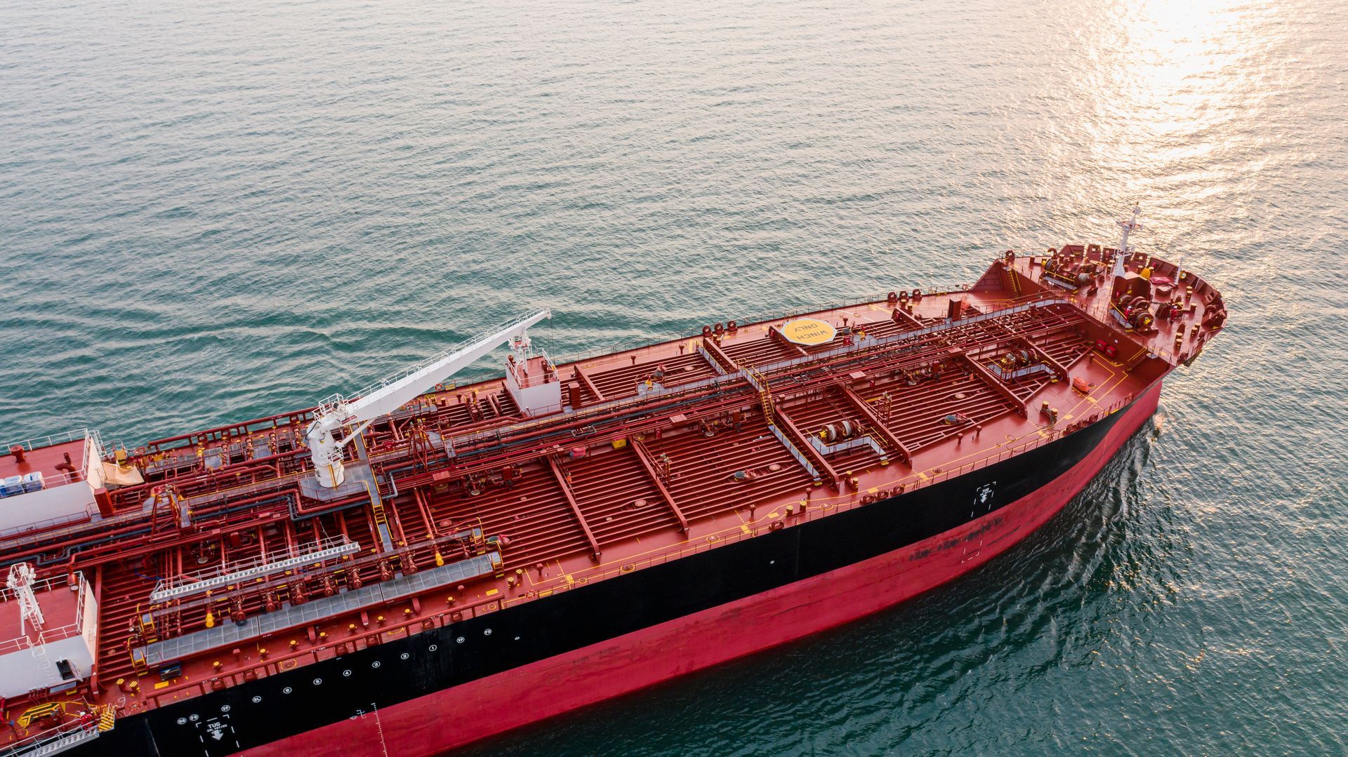 Large red and black oil tanker on the ocean, sun reflecting off the water.