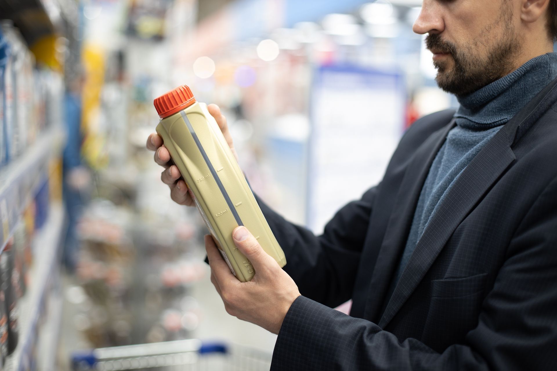 Man in a blazer examines motor oil bottle in a store aisle, selecting a product.
