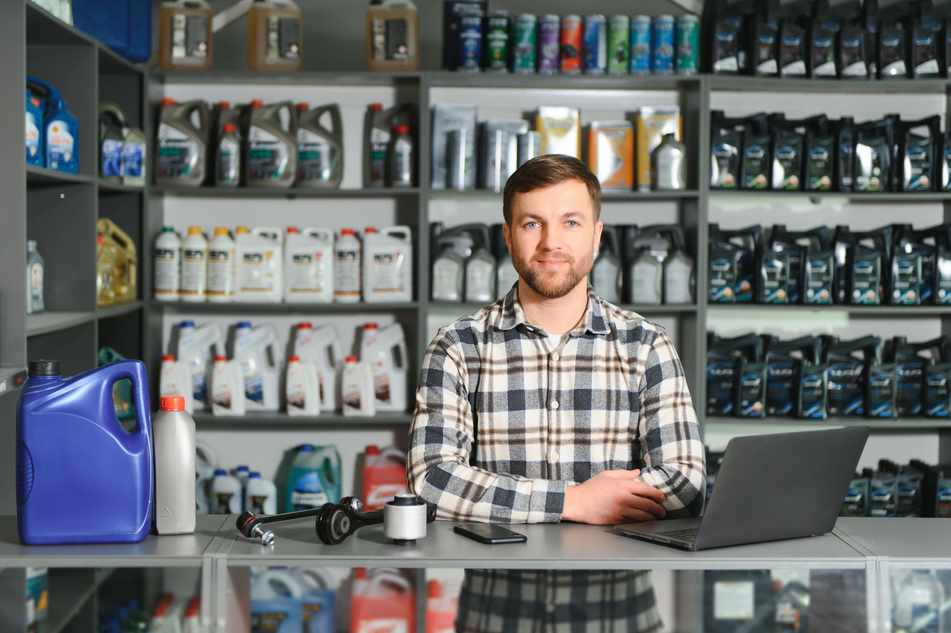 Man in plaid shirt behind a counter with laptop, automotive parts and shelves of products in a store.