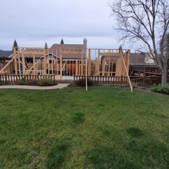 Wood-framed house under construction behind a grassy yard, with bare trees and a cloudy sky