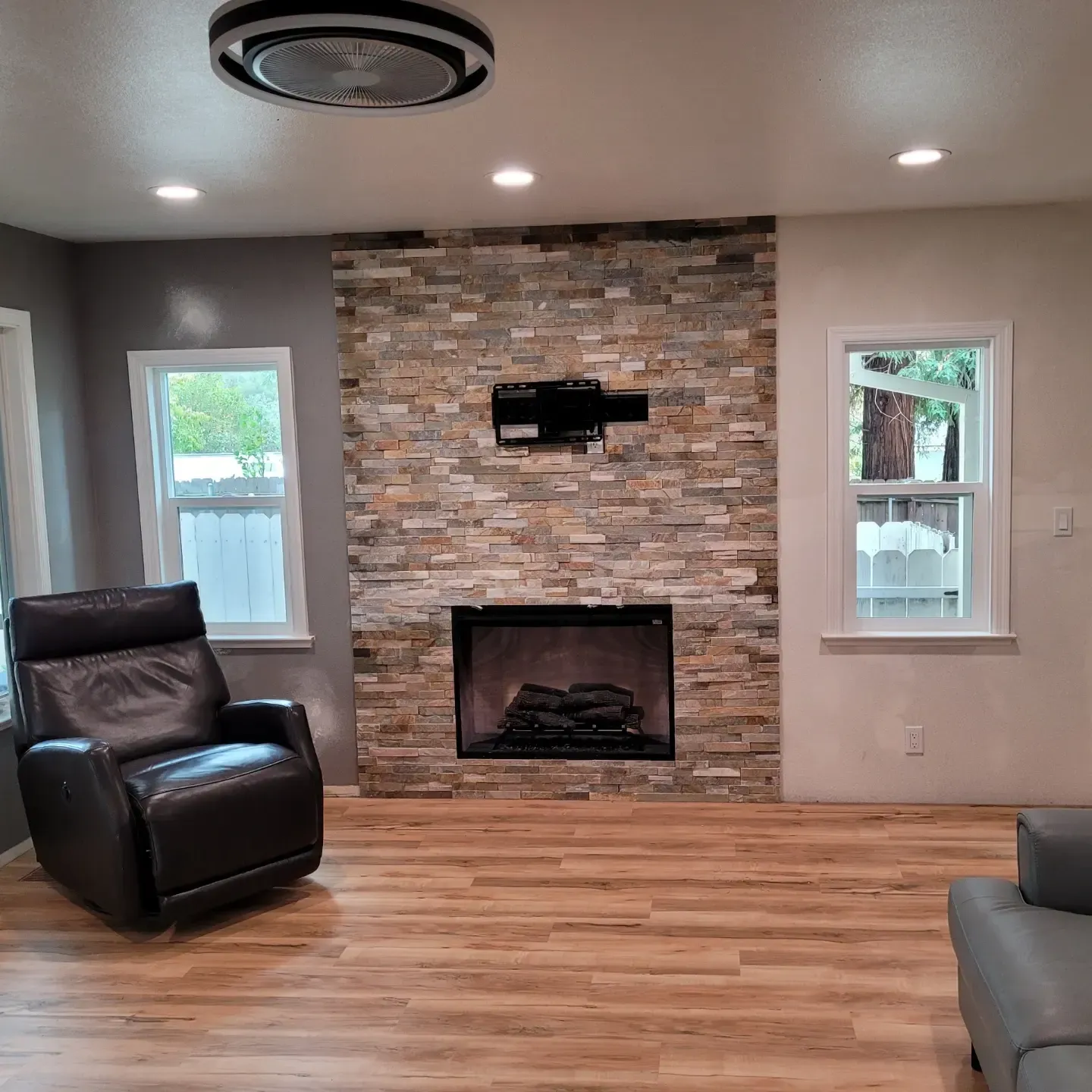 Living room with stone fireplace, wood floor, leather chair, and two windows.