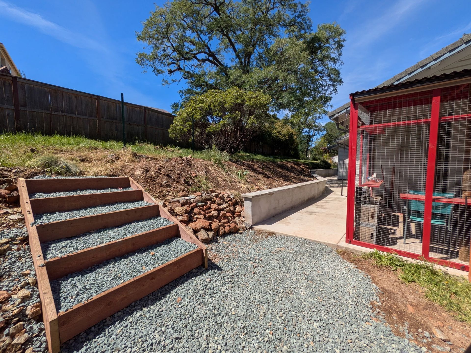 Backyard with gravel path, wooden steps, retaining wall, and red-framed screened porch under a blue sky