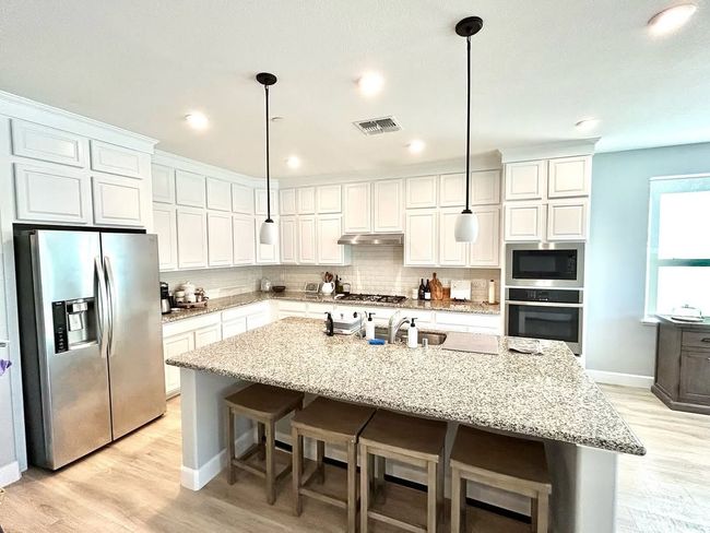 Bright white kitchen with granite island, stainless fridge, double ovens, and pendant lights