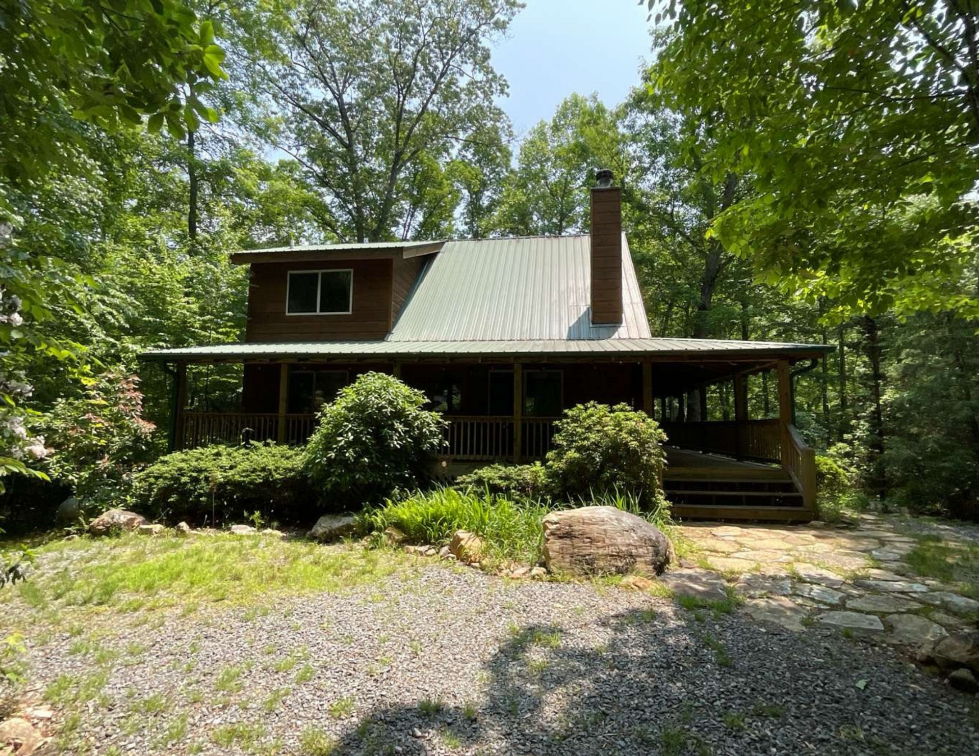 Cabin with a green roof, porch, and stone pathway surrounded by trees.