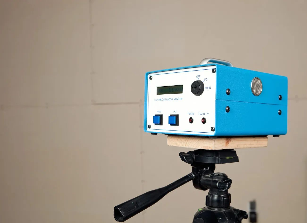 A blue electronic device on a tripod, against a tan wall. Has a screen, knobs, and buttons.