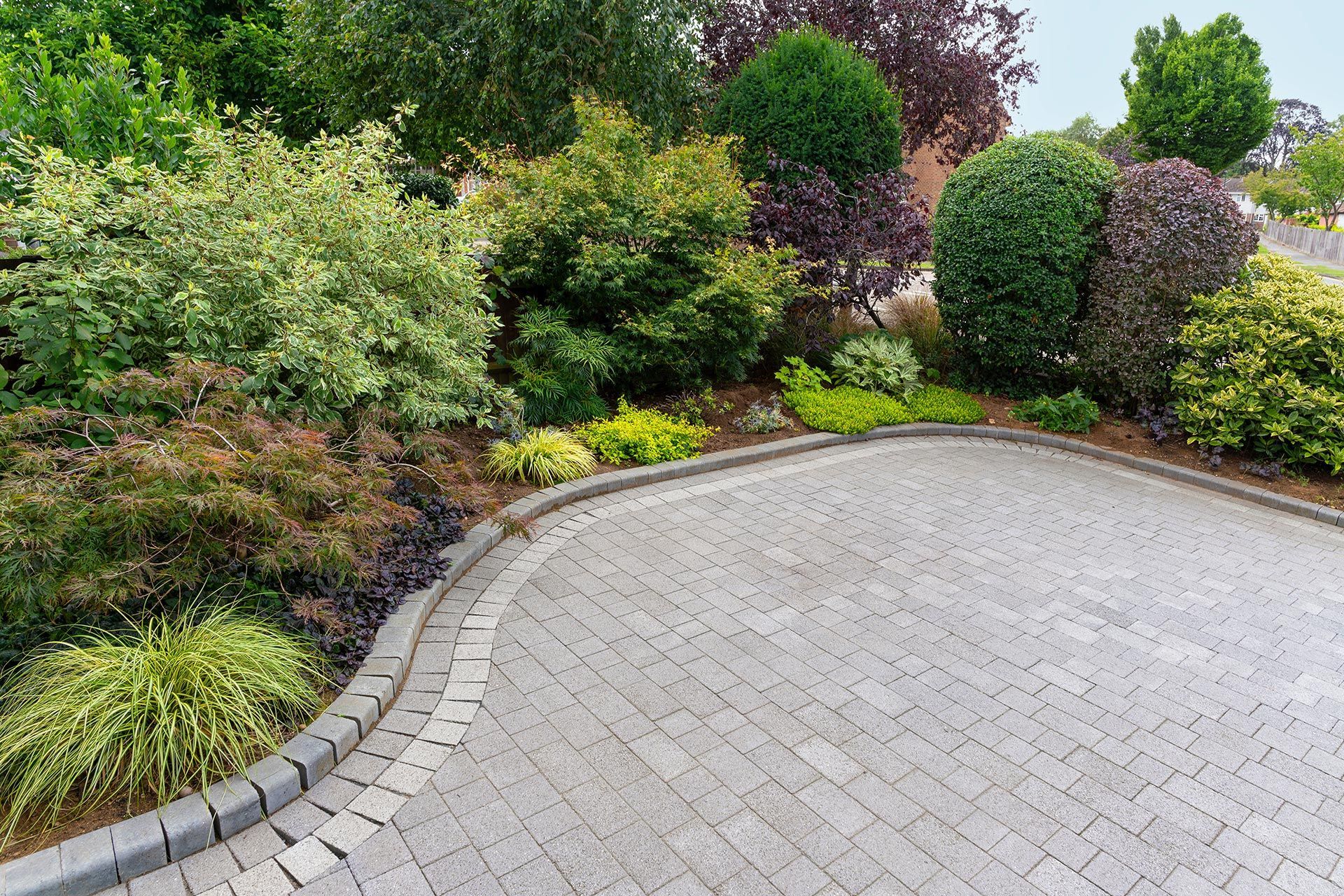 Curved stone driveway bordered by lush green shrubs and colorful garden plants