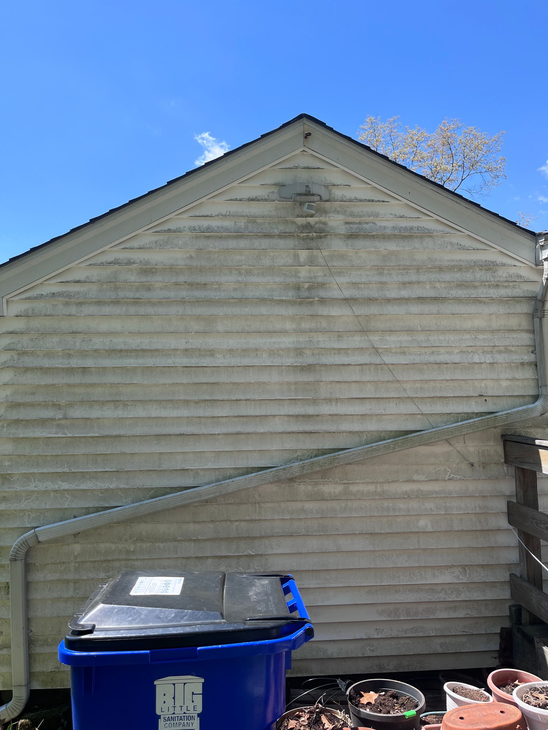 Weathered house siding with a blue trash bin and a flower pot in front under a clear blue sky
