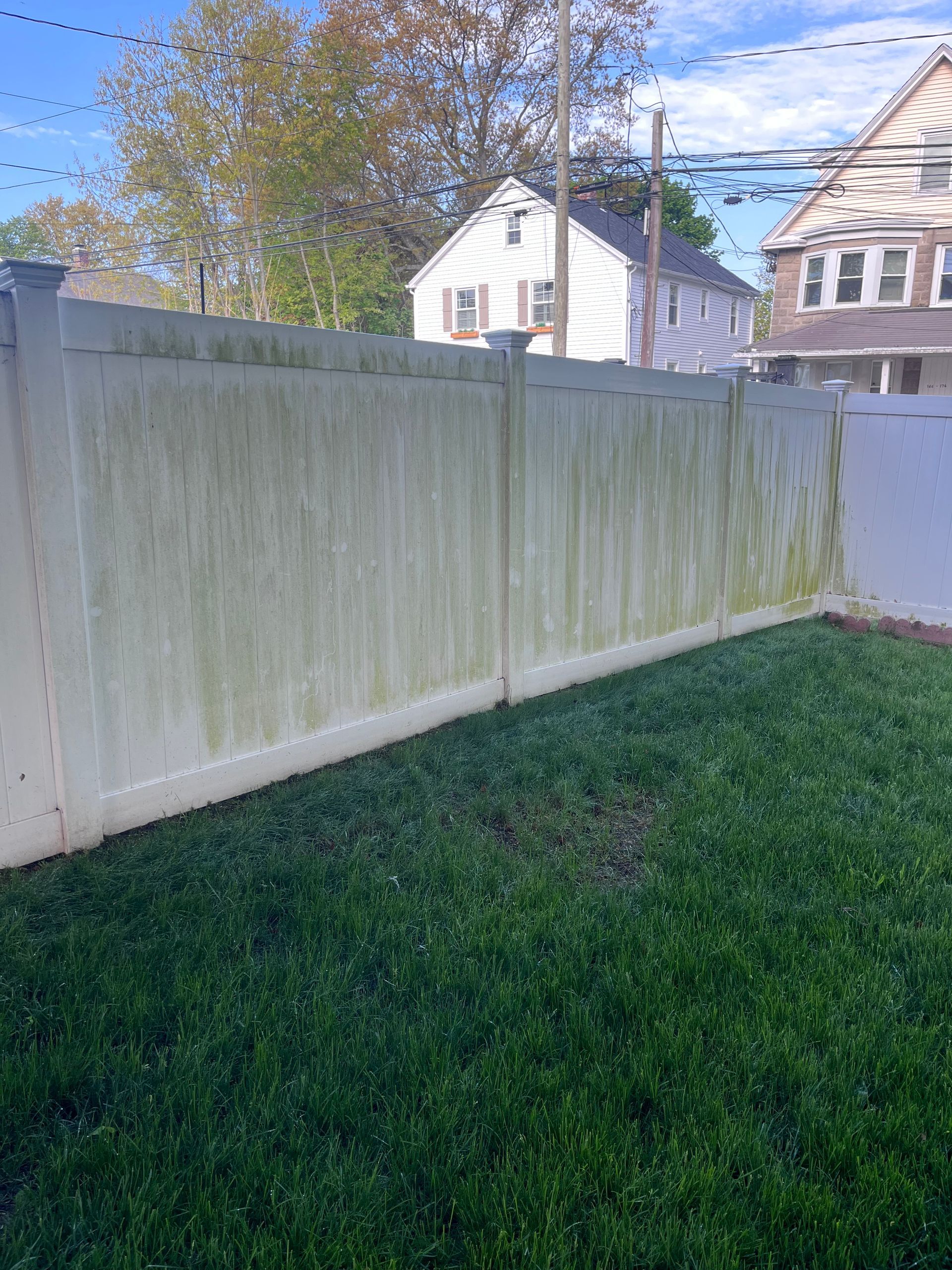 White wooden fence beside a green lawn, with houses and a tree in the background.