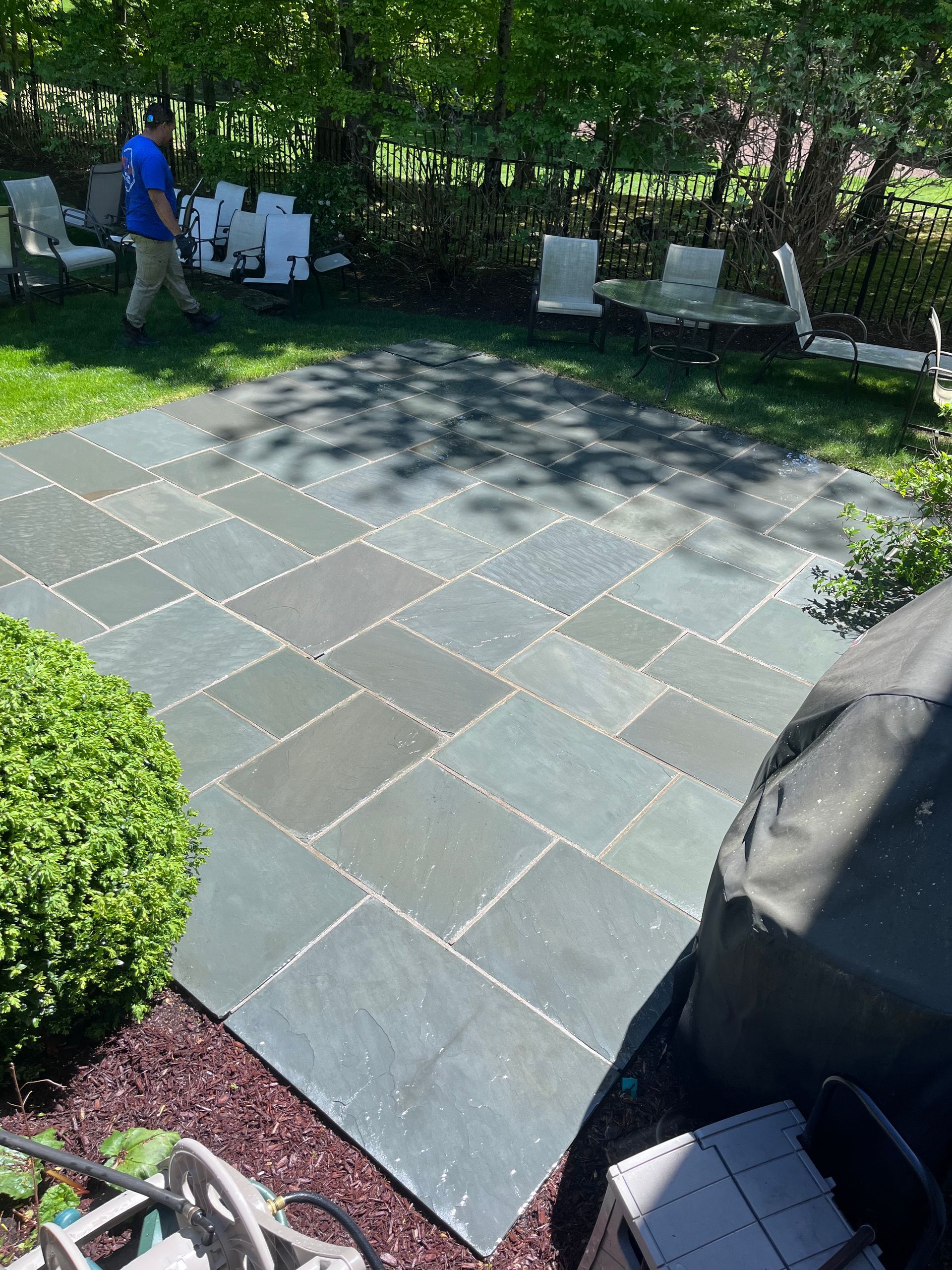 Backyard stone patio with chairs and a person in a blue shirt near a shed, dappled in shade.