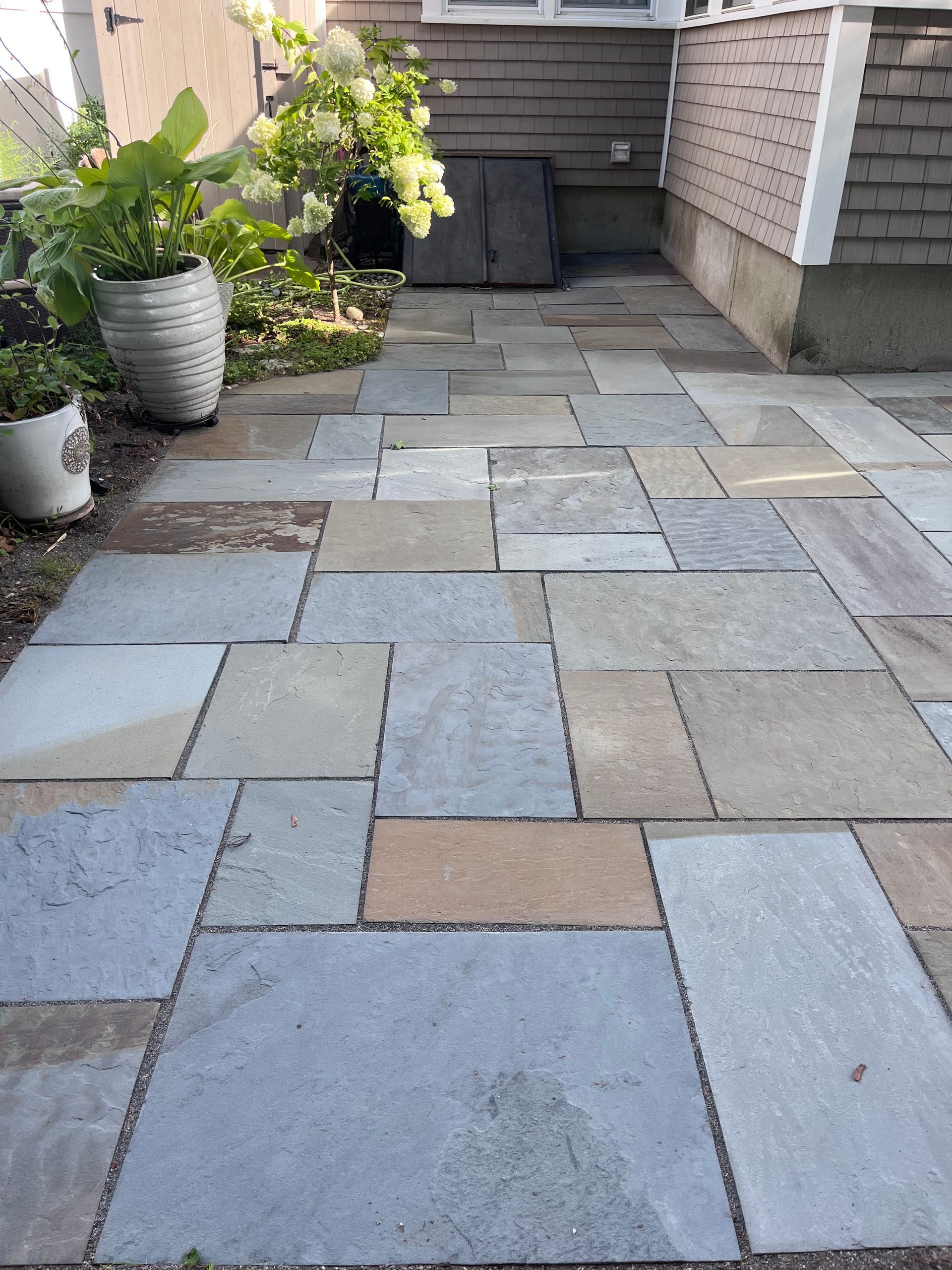 Stone paver patio walkway beside a house, with potted plants along the left edge.
