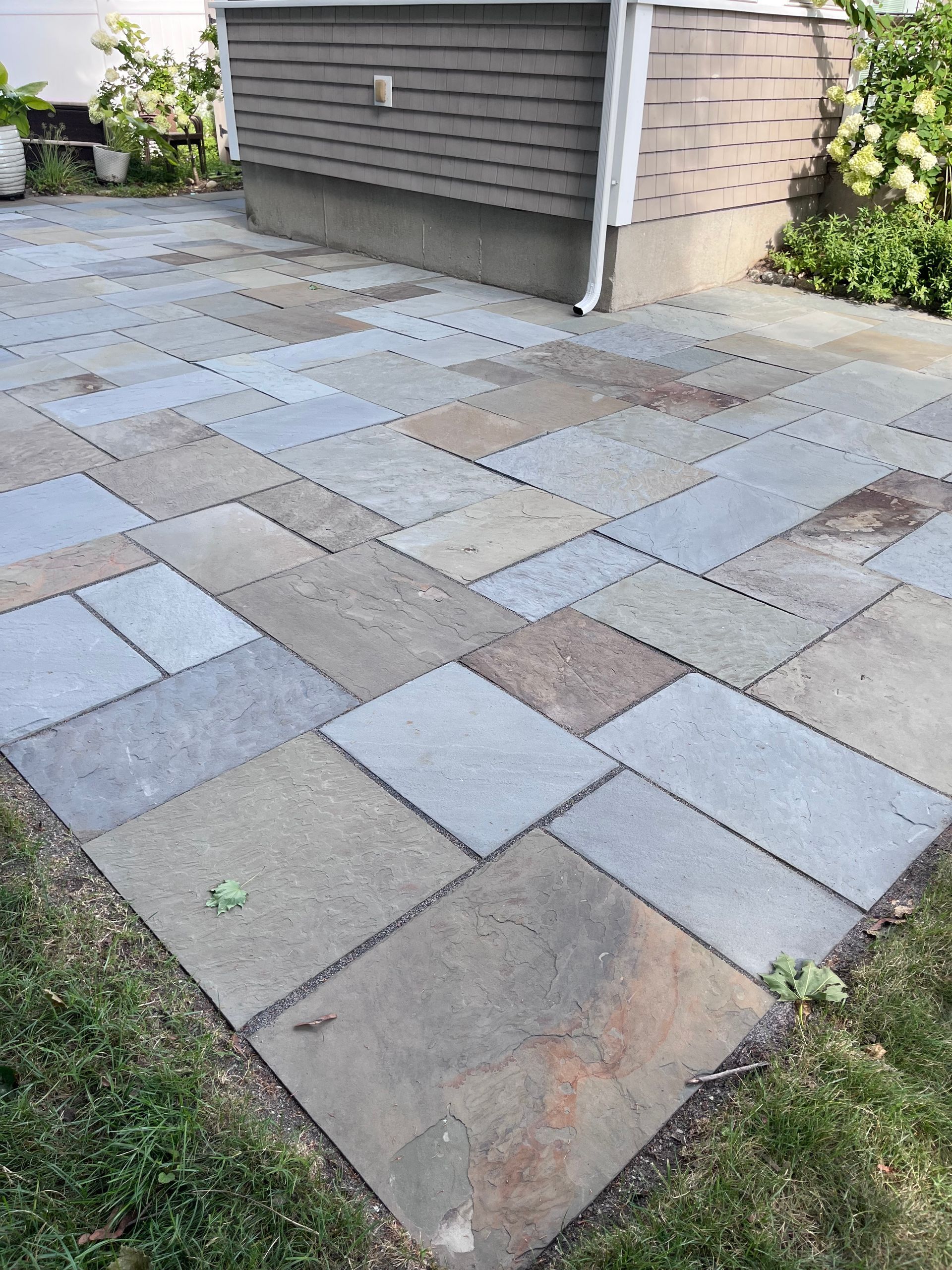 Stone patio with multicolored pavers beside a house and garage wall, bordered by grass and shrubs