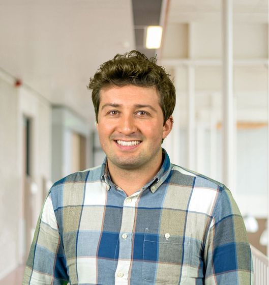 A person with dark hair smiling while wearing a blue and white plaid collared shirt in a bright office hallway.