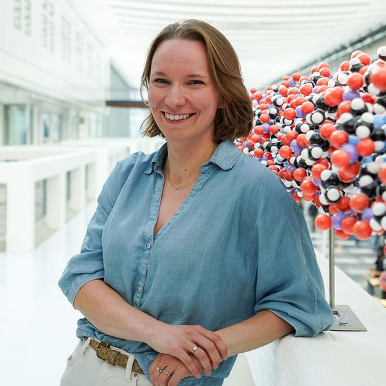 A person in a beige blazer and dark top leans against a large, colorful molecular model structure in a bright hallway.