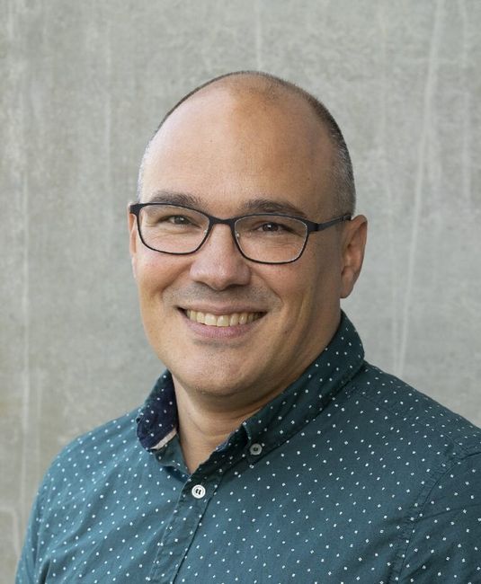 A person wearing glasses and a polka-dot button-up shirt smiles while standing in front of a plain grey background.