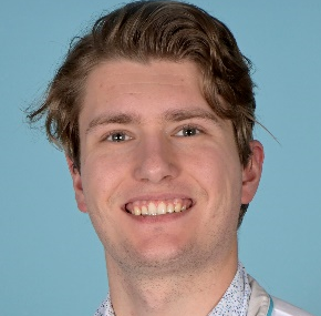 A person with wavy brown hair smiling at the camera, wearing a white patterned shirt against a light blue background.