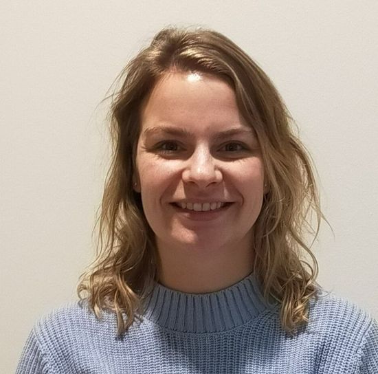 A person with shoulder-length blonde hair and a friendly smile, wearing a light blue ribbed sweater against a white wall.