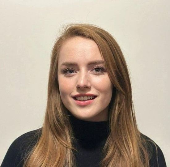 A person with long blonde hair and a neutral expression, wearing a black mock-neck top against a plain light background.