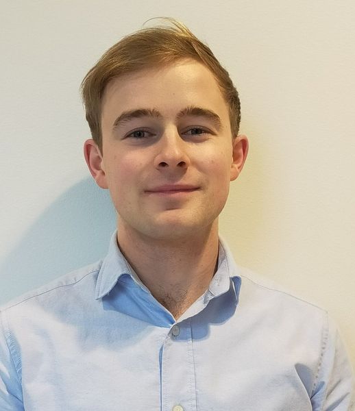 A person with light brown hair wearing a light blue button-down shirt, standing against a plain, off-white wall.