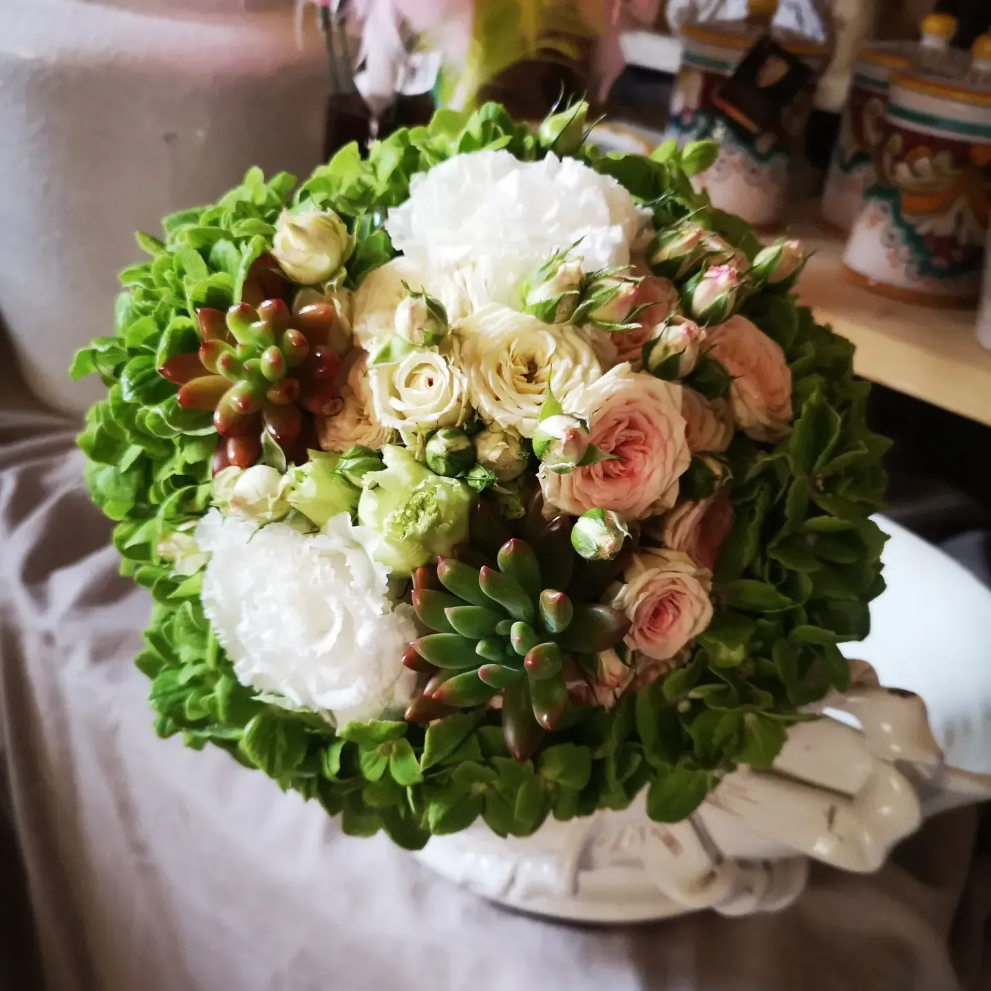 Un bouquet da sposa composto da rose bianche e rosa cipria, ortensie verdi e piante succulente in un vaso bianco su un tavolo.