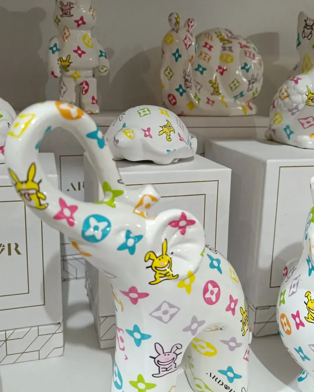 Statuette di animali in ceramica bianca decorate con colorati loghi in stile Louis Vuitton e personaggi dei cartoni animati, disposte su degli scaffali.