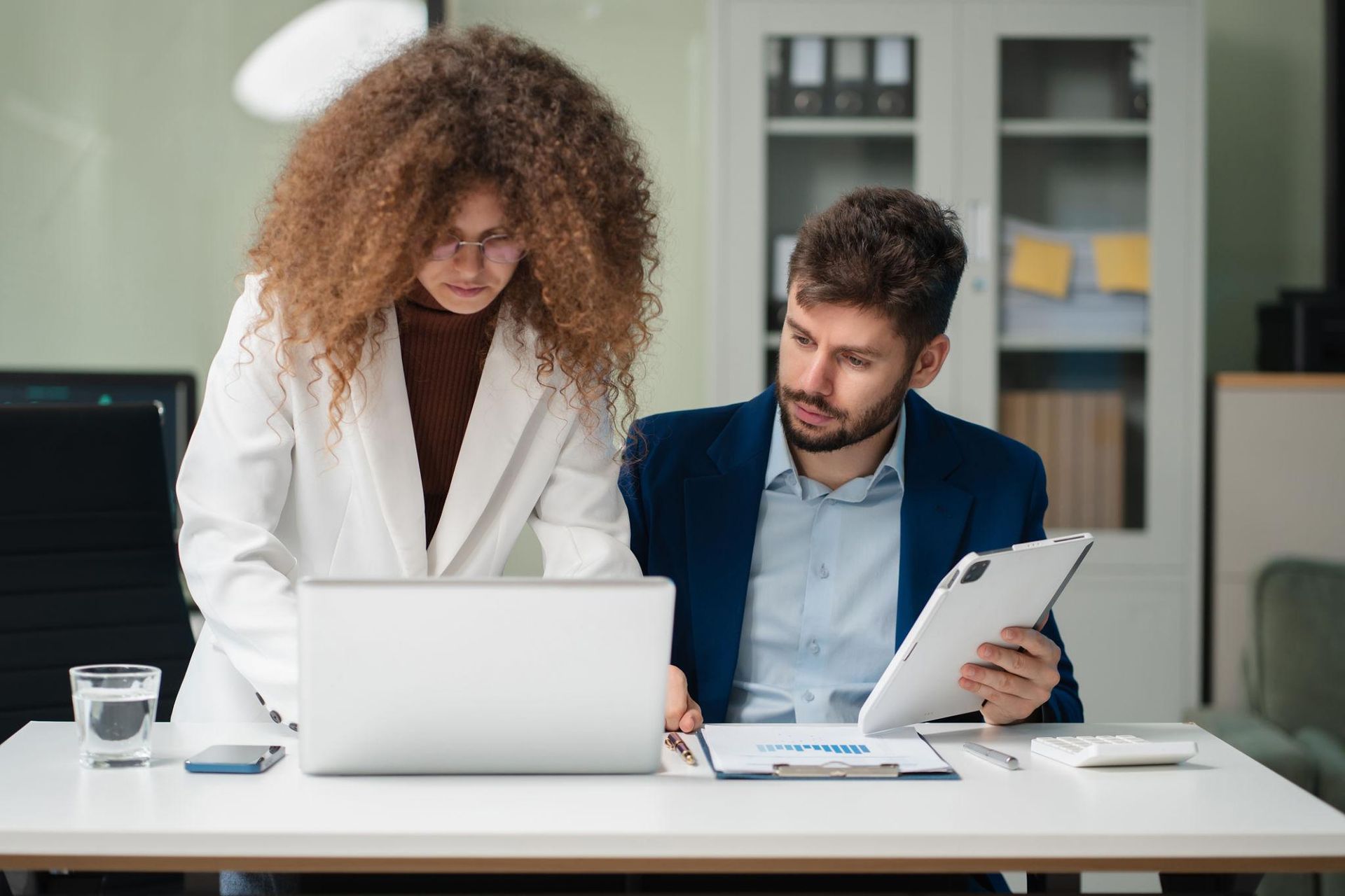 Two coworkers in an office reviewing documents beside a laptop and tablet