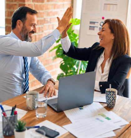 Two coworkers in a meeting high-five across a desk with a laptop and papers.