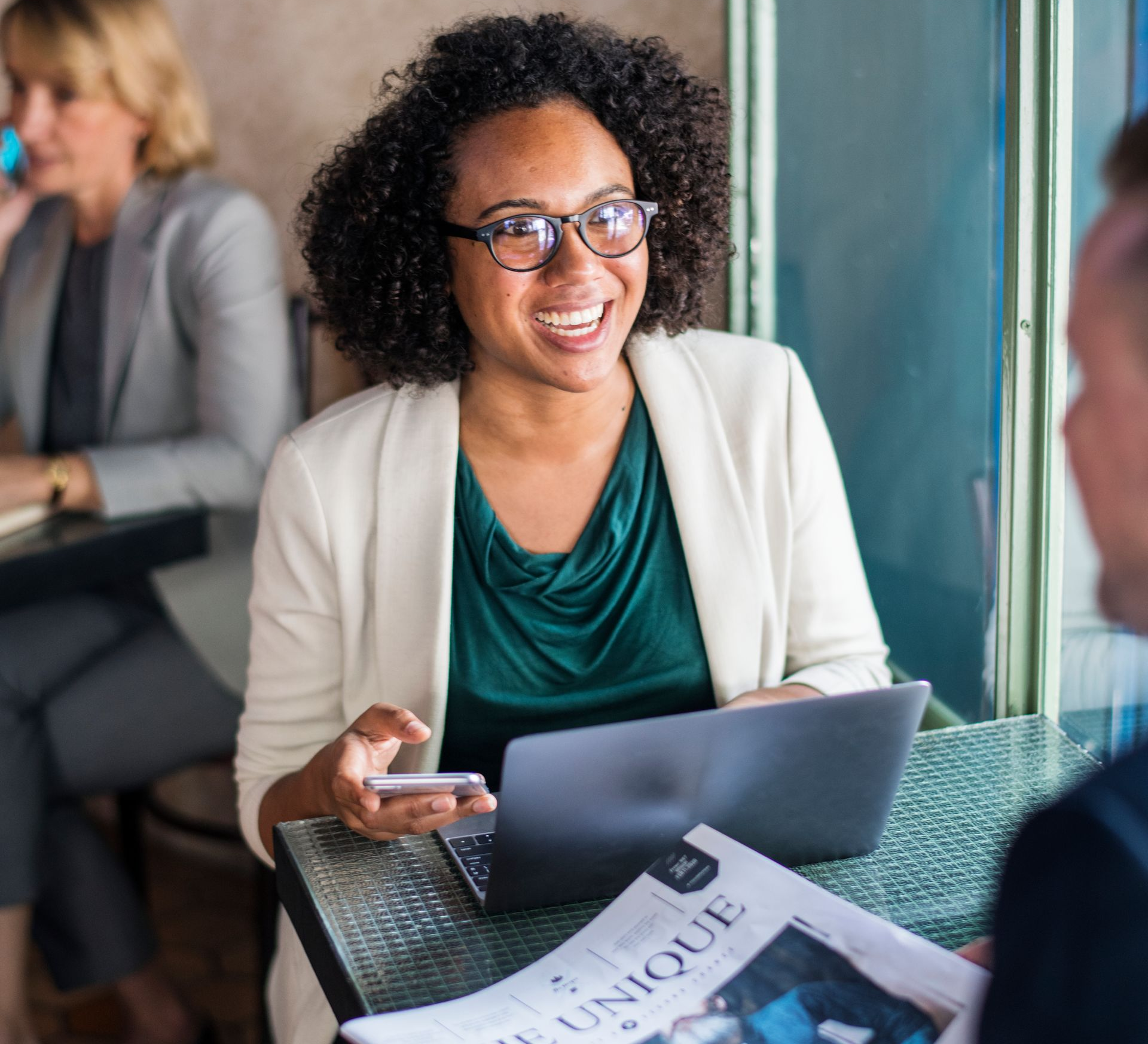 Smiling person with laptop and phone at a café table, papers in front, meeting with someone across from them