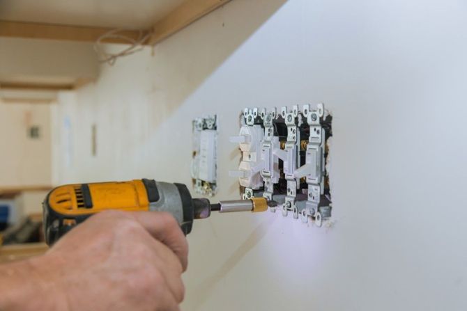 Hand holding a drill while removing an electrical outlet cover on a wall