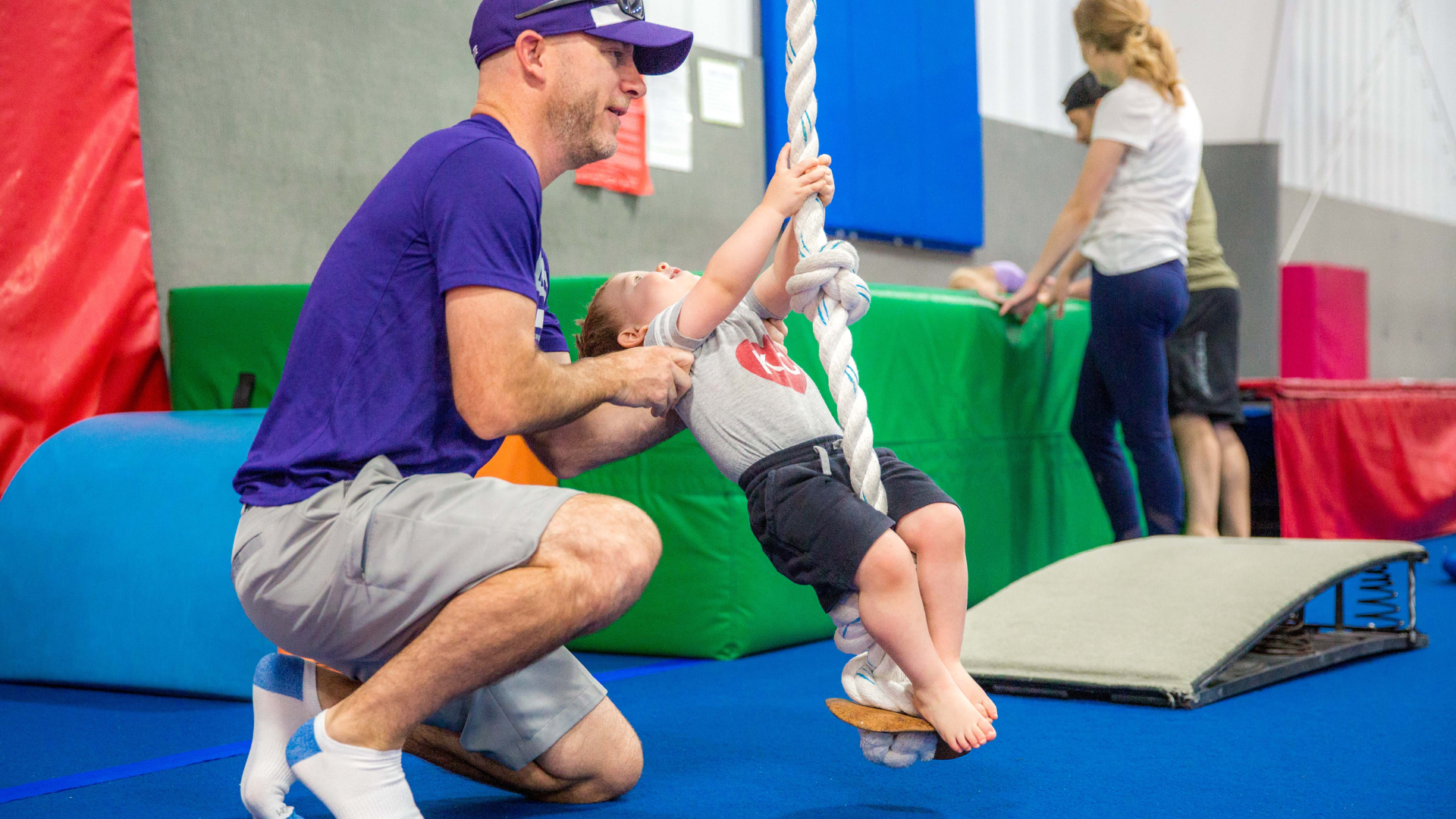 A dad in a purple shirt is helping his child swing on a rope.