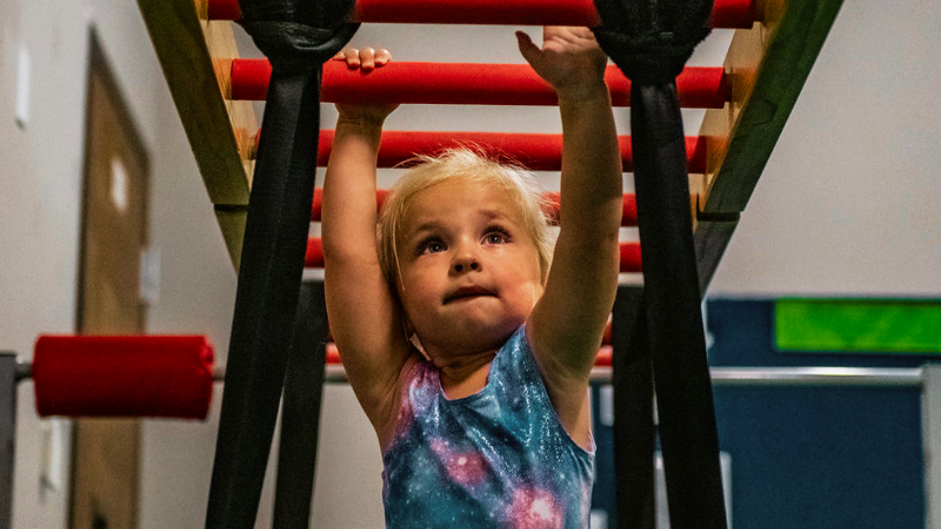 A preschool gymnast is reaching across the bar in a blue and pink leotard