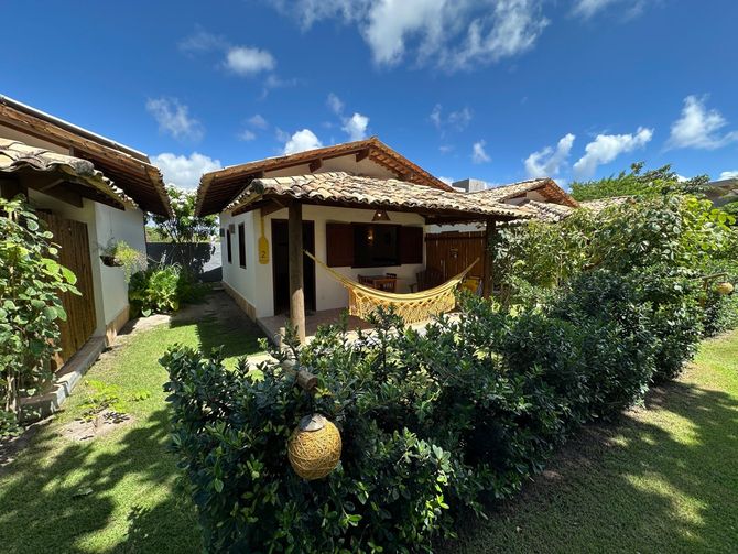Bangalô tropical com telhado de telha, paredes brancas e um exuberante jardim verde sob um céu azul brilhante.