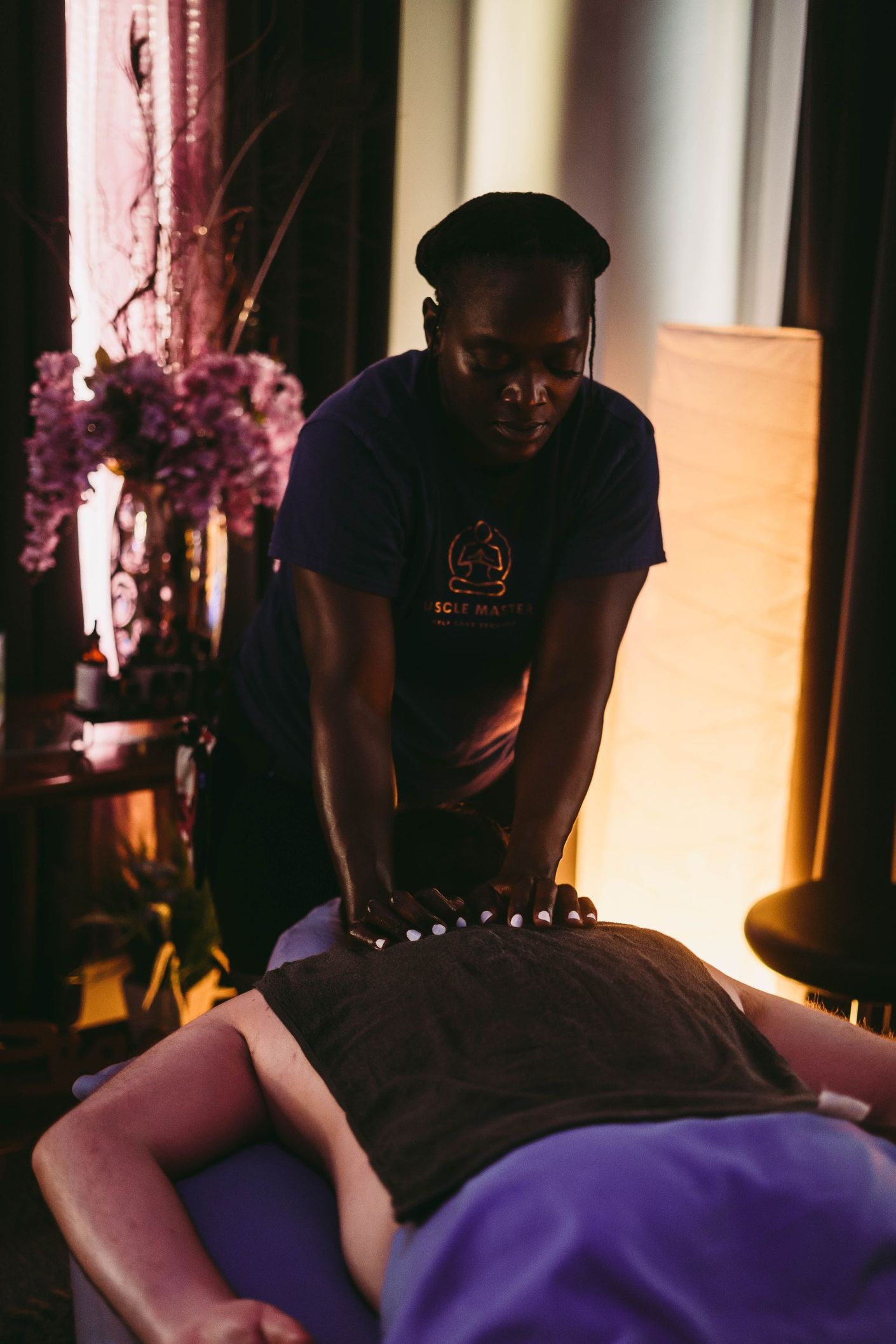 A person performs a back massage on a client lying on a treatment table in a softly lit, warm-toned room.