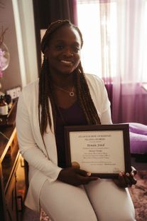 A smiling person in white clothing holds a framed certificate in a softly lit indoor room.