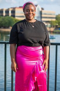 A person in a black t-shirt and bright pink ruffled skirt stands smiling by a railing with a river and building behind.