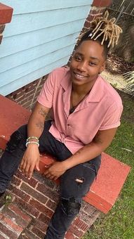 A person with styled blonde hair, wearing a pink shirt and ripped black jeans, sitting on a red brick ledge.