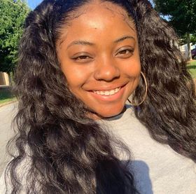 A smiling person with long, dark, wavy hair and gold hoop earrings posing outdoors in sunlight.