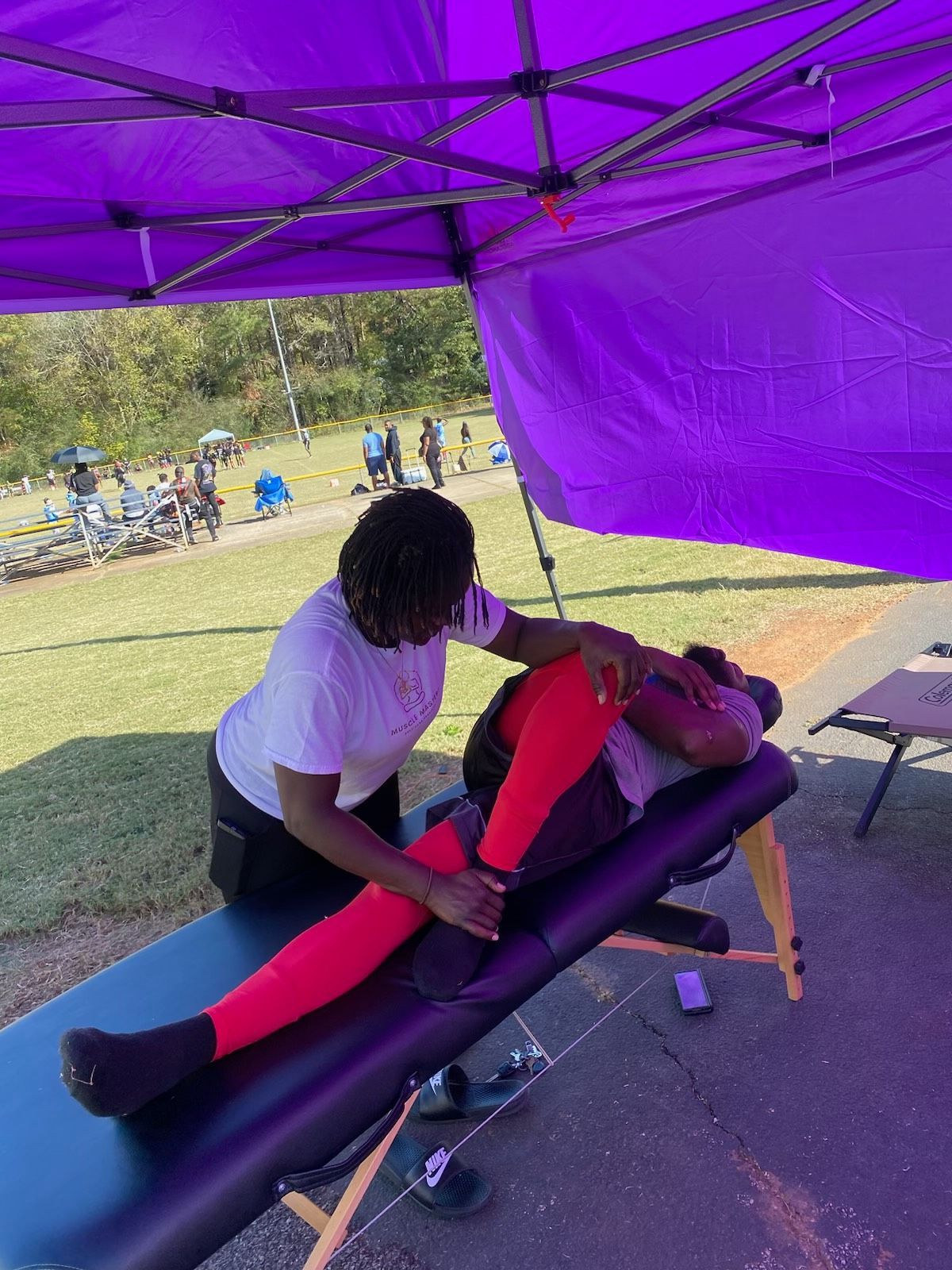 A practitioner performs a leg stretch on a person lying on a massage table under a purple canopy at an outdoor event.