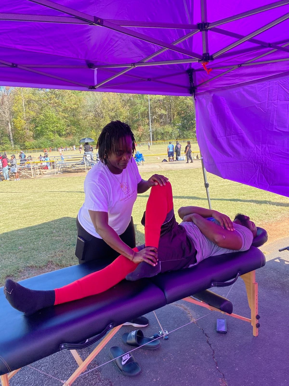 A practitioner performs a leg stretch on a person lying on a massage table under a purple canopy at an outdoor event.