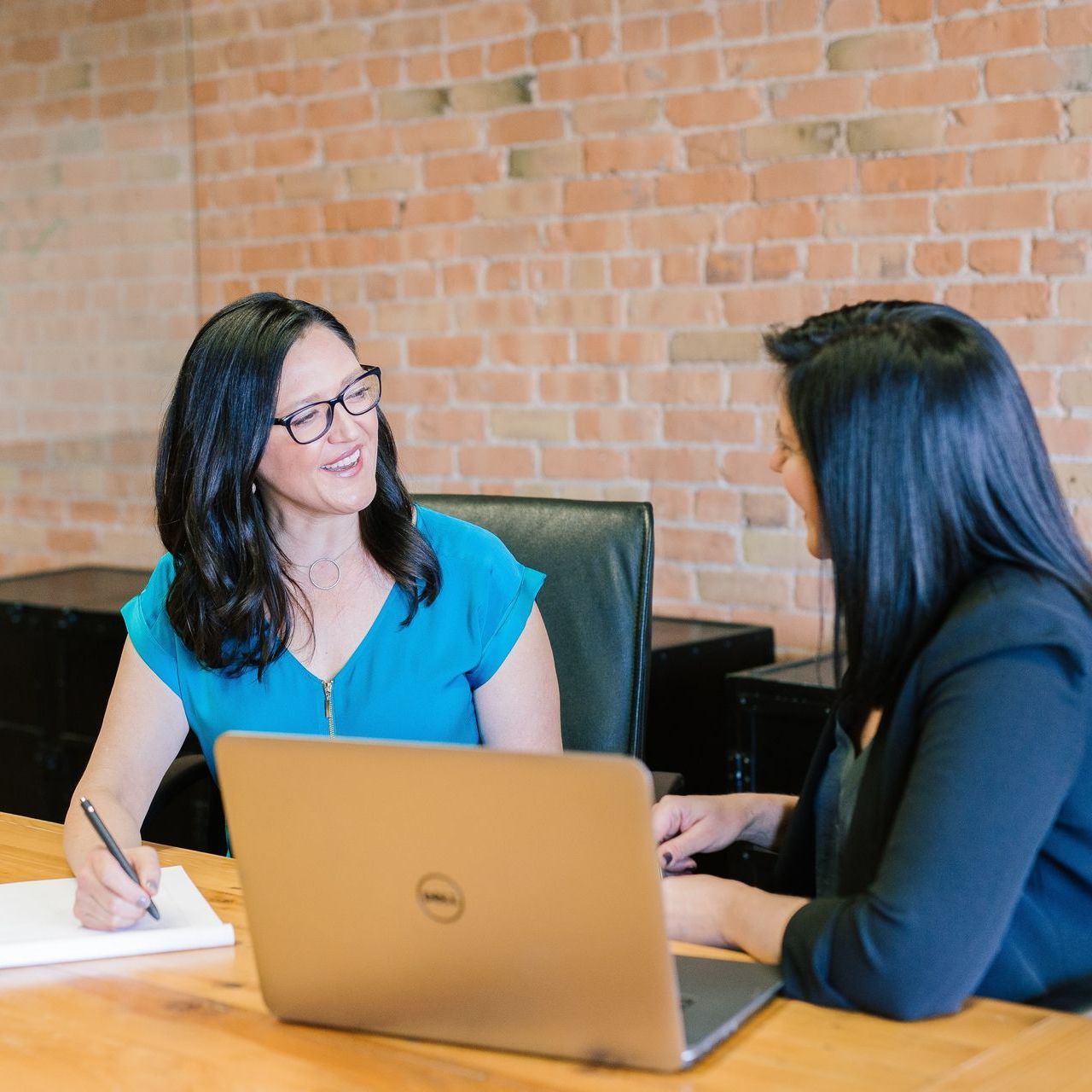 Two women at a table in an office; one writing, one on laptop; brick wall background.