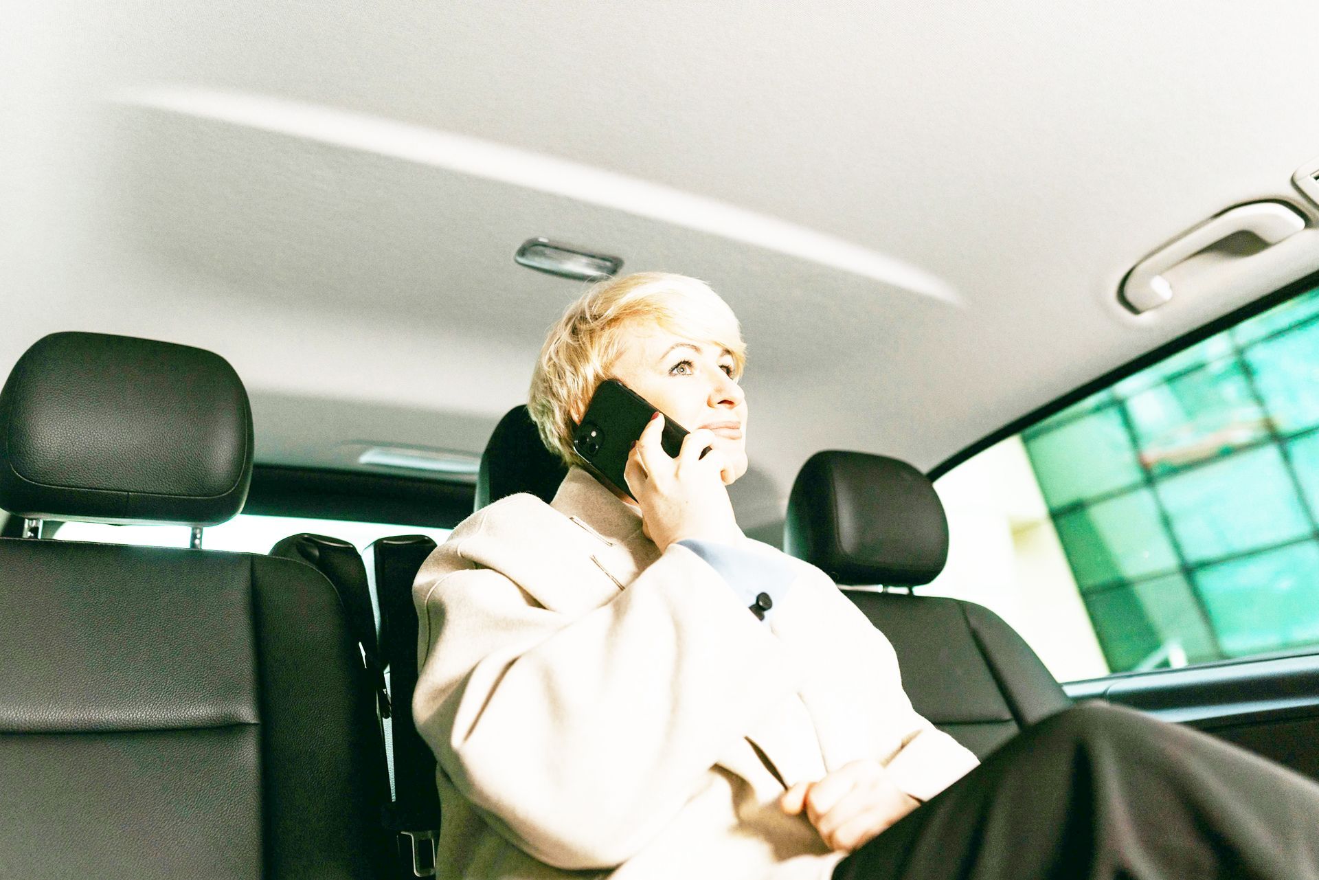 Woman in car talking on a phone, light clothing, black seats, bright window.