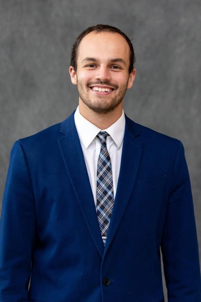 Smiling man in a blue suit and patterned tie against a gray studio backdrop