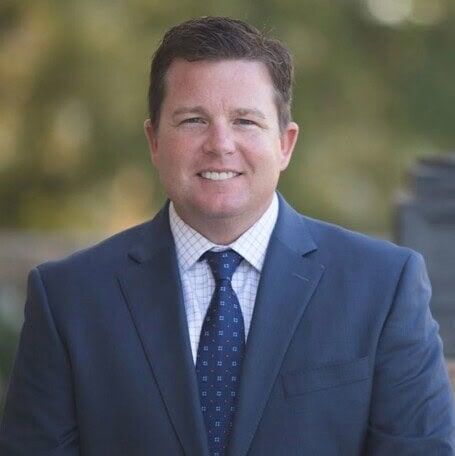 Smiling man in a blue suit and tie outdoors, with a blurred background.