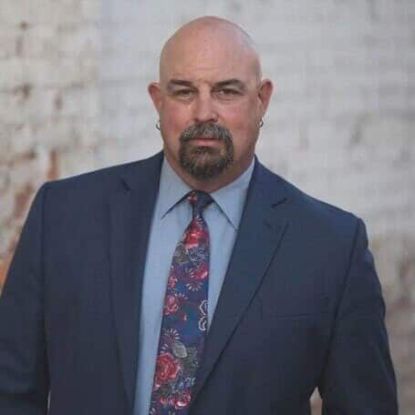 Portrait of a bald man in a navy suit and floral tie against a light stone wall