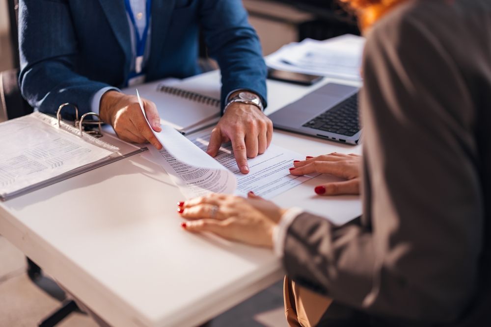 Two business people at a desk review documents. One points to a paper.