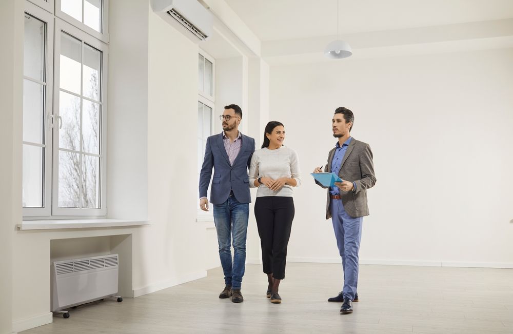 Three people in an empty room, one showing property to a couple. White walls, large window.