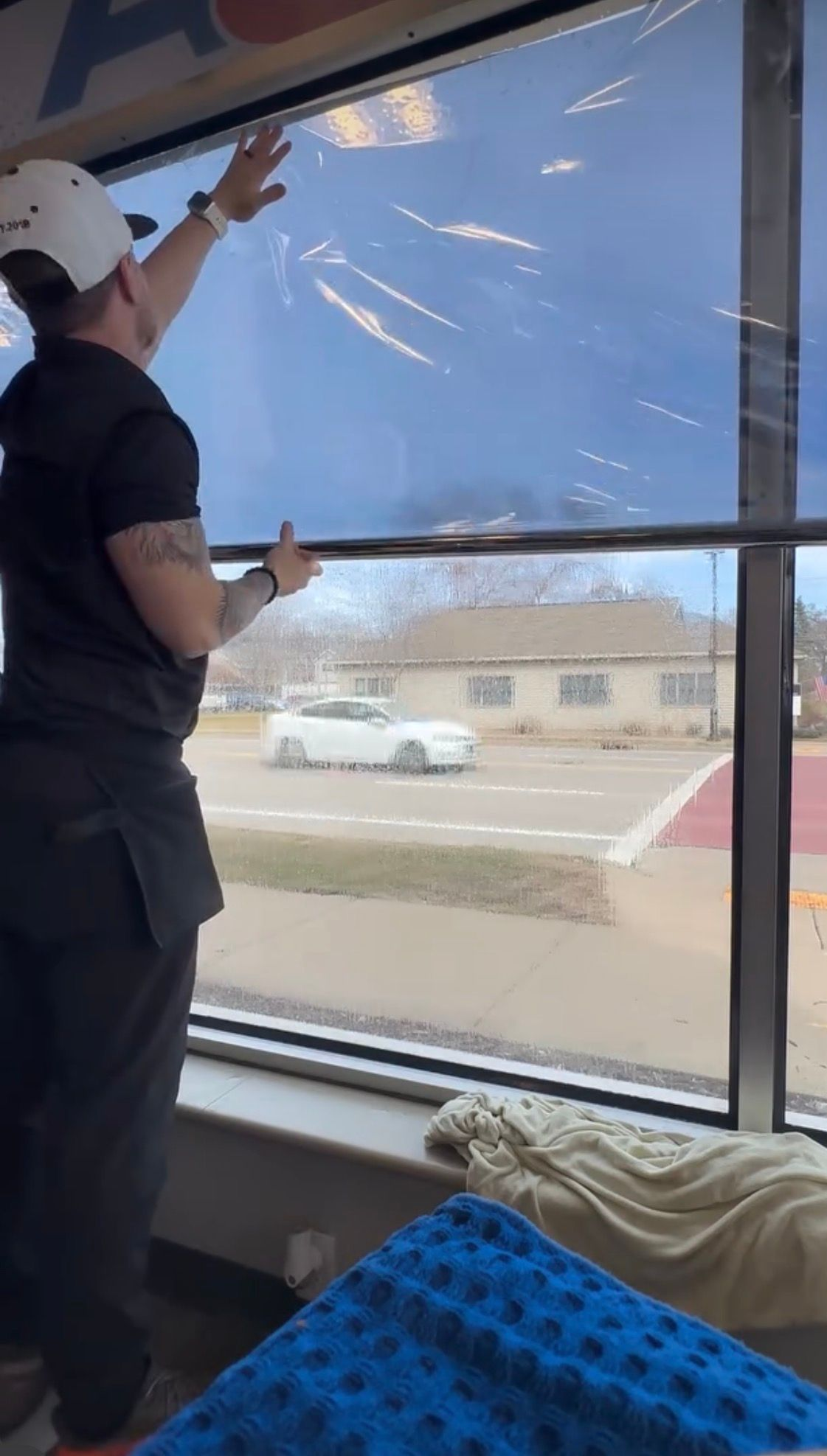 A person in a black shirt and white cap installs tinted window film on a large glass window overlooking a street.