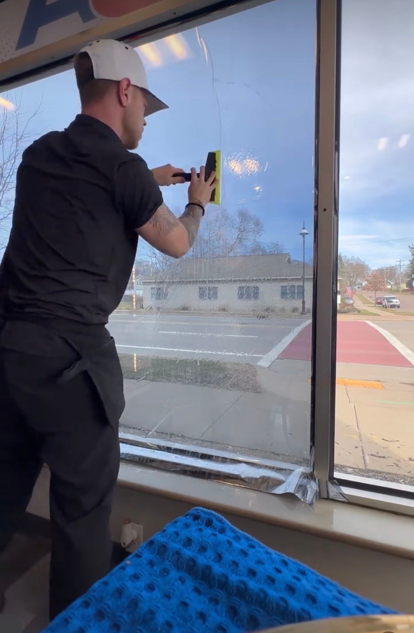 A person wearing a hat and black uniform cleans a large store window with a yellow scrubbing tool.